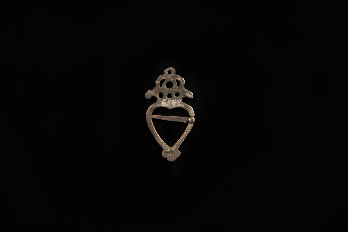 En hjärtformat bröstsölja av silver (?), med genombruten krona överst. Fästanordning av en nål. Nedtill treklöverformad avslutning. Troligen från Halland.