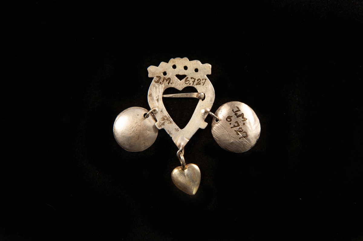 Ett hjärtformat spänne av förgyllt silver. Graverad dekor av punkter m.m. Två skålar och ett hjärta av silver hänger från spännet. Rester efter gravyr på en av skålarna visar att återanvänt material har använts.