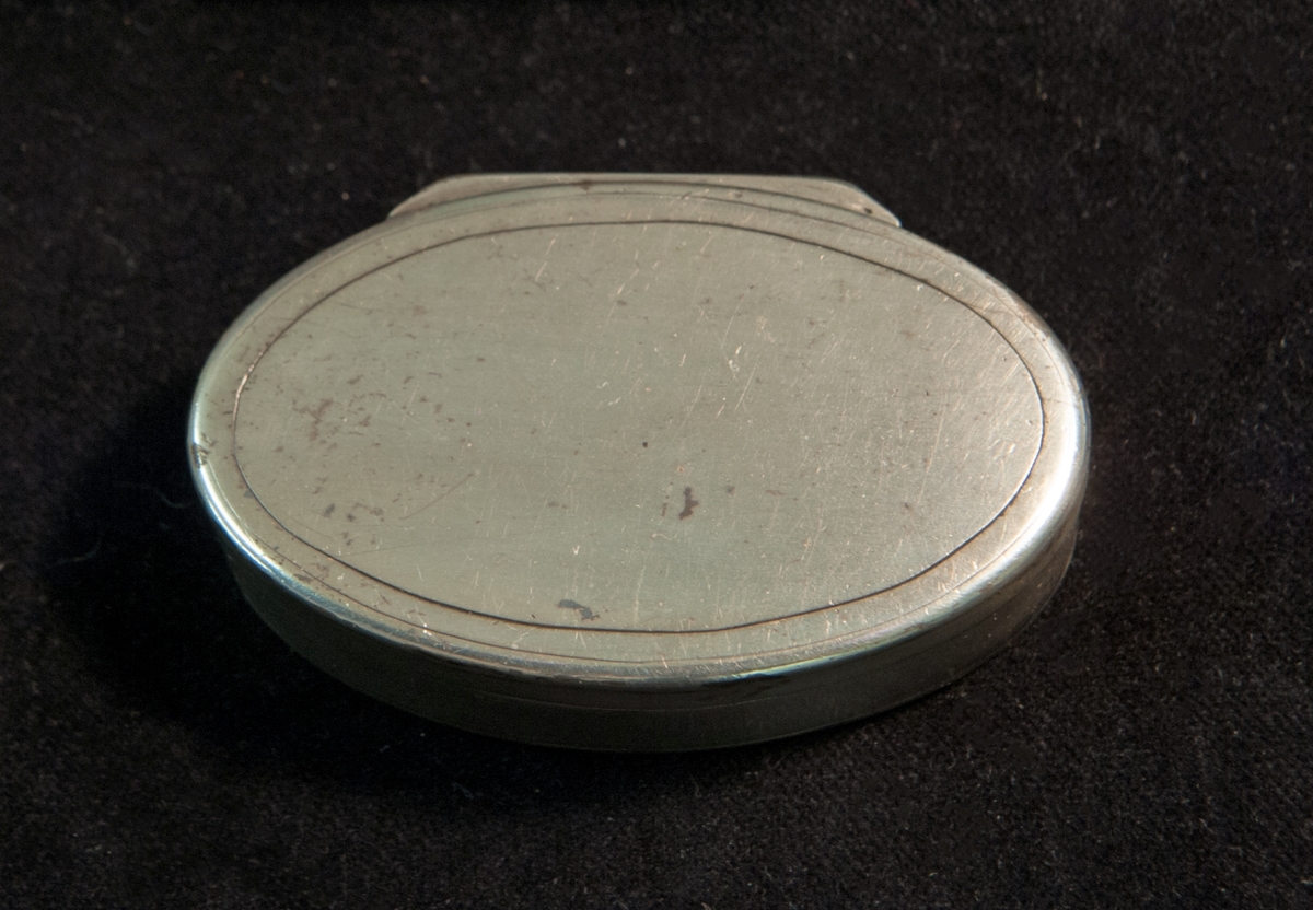 En oval dosa av silver. Slät modell med randdekor. I botten graverade ägarinitialer i spegel monogram: "JH ". Invändigt förgylld. Stämplad invändigt i botten.