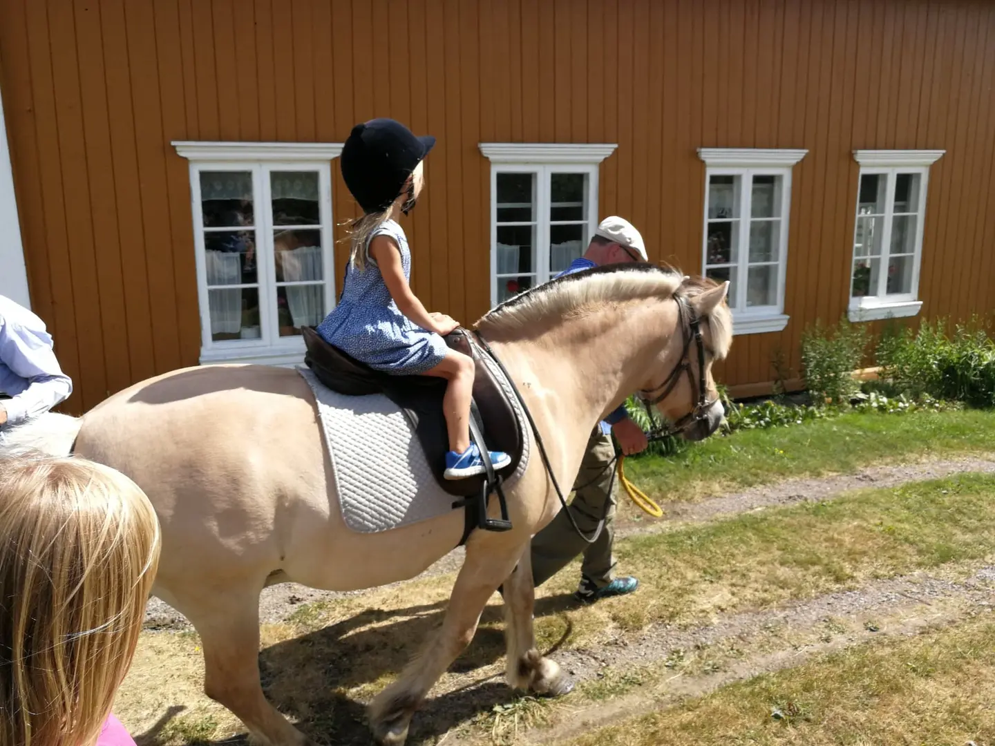 Ridetur på fjordhest under Middelalderdagen på Almenninga. Ei ung jente rir forbi Austun. Austun er sennepsgul og har hvite vinduskarmer. Sommerdag.