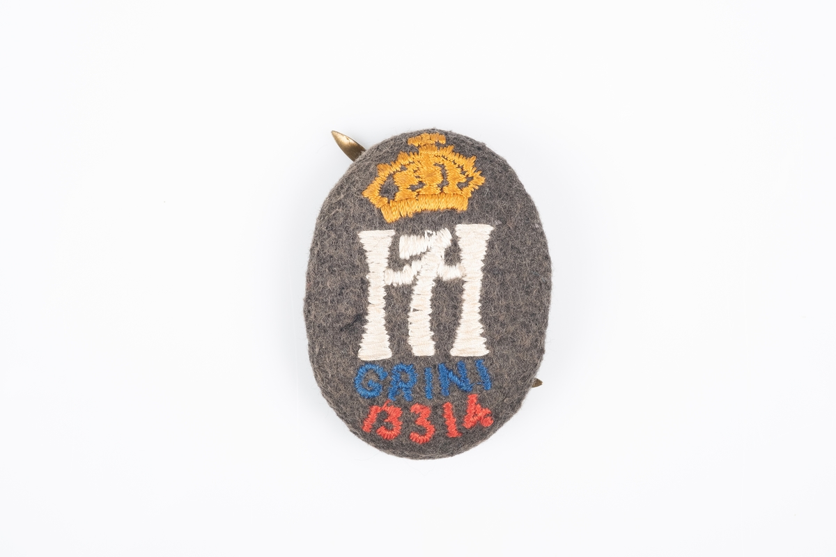 En medaljong i tekstil med to splittbinders på baksiden. Framsiden har brodering av "H7" med krone, "Grini" og fangenrummer.