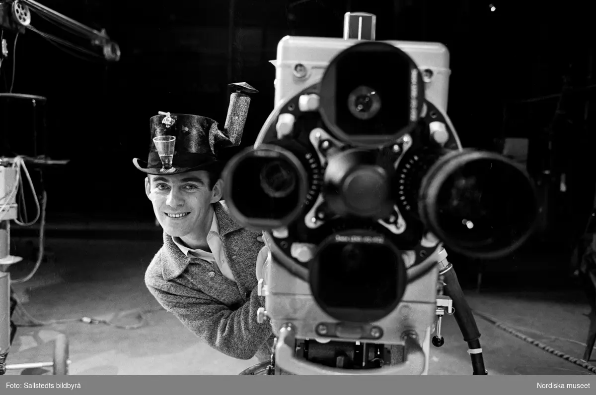 Foto från TV-inspelning på Cirkus med Gunnar Wiklund. Troligen inspelning av "Gunnar Wiklund show". Gunnar Wiklund står vid en tv-kamera. På huvudet har han en hög hatt med en inbyggd kran, skorsten och ett snapsglas på brättet.