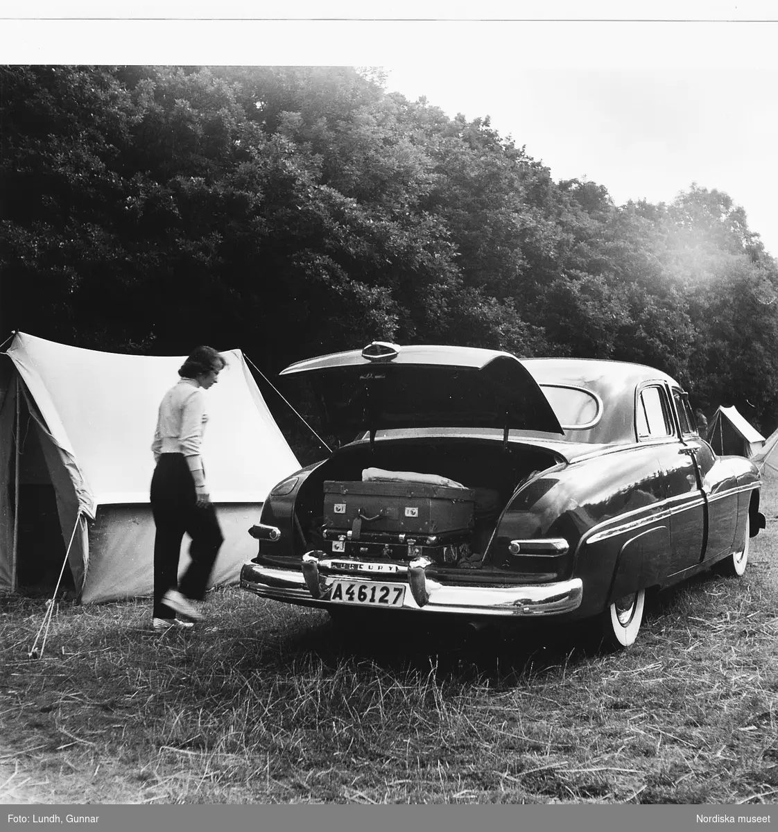 En kvinna står bredvid en Mercury årsmodell 1950. Bagageluckan är öppen och det ligger resväskor i bagaget. Bredvid bilen står ett tält. Bilden tagen på Mölle camping år 1952.
