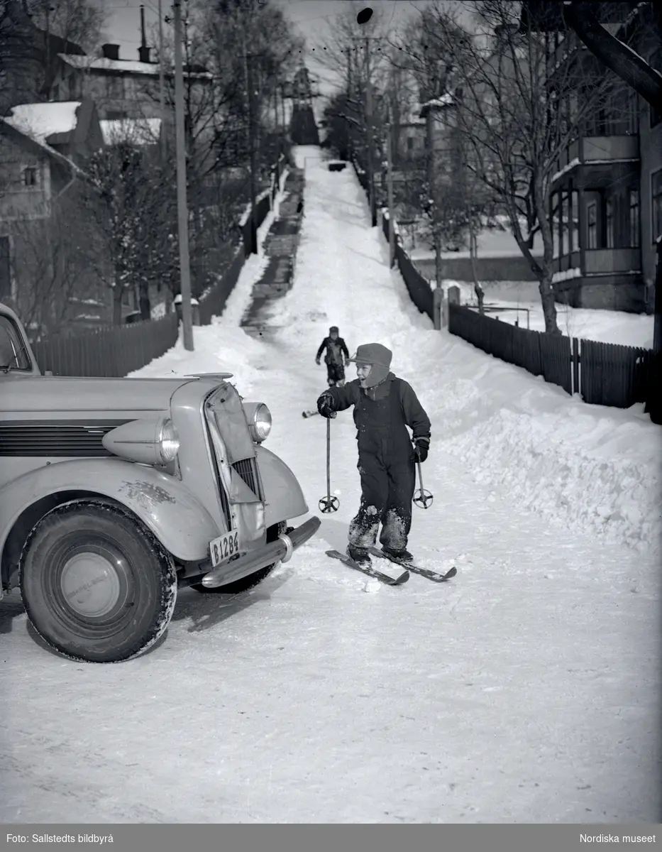 Enligt påskrift "Trafikfälla i Solna" år 1947. Vy över en gata i ett villaområde. Ett barn på skidor står framför en bil. Marken är snötäckt.