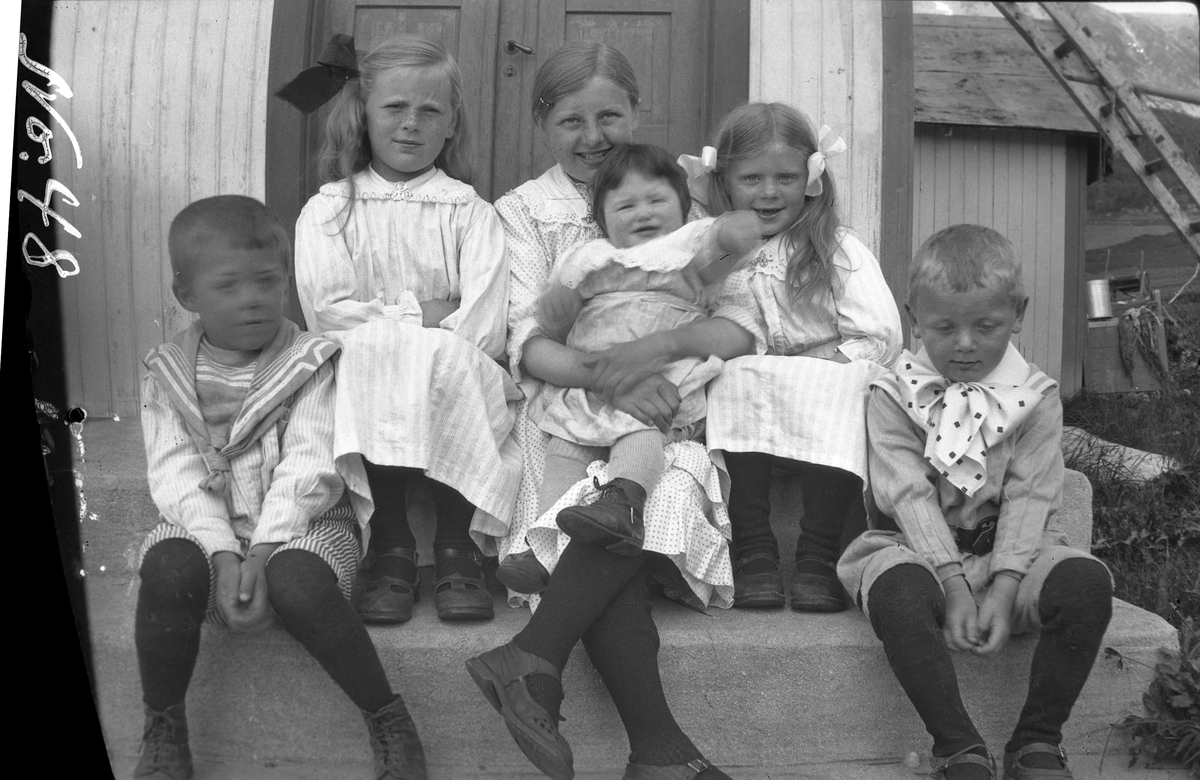 Gruppeportrett av seks barn, fotografert sittende på en trapp utendørs.