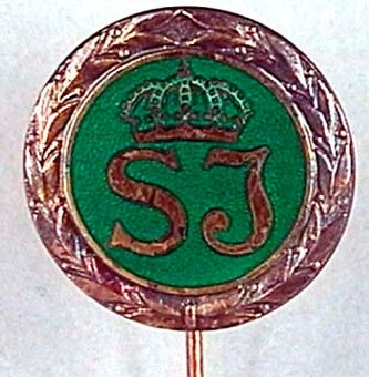 Utmärkelsetecken, runt med nål för fastsättning i kavajslag. Märket är av silver med SJ:s logotyp på grön emaljerad bottenm inom en lagerkrans.