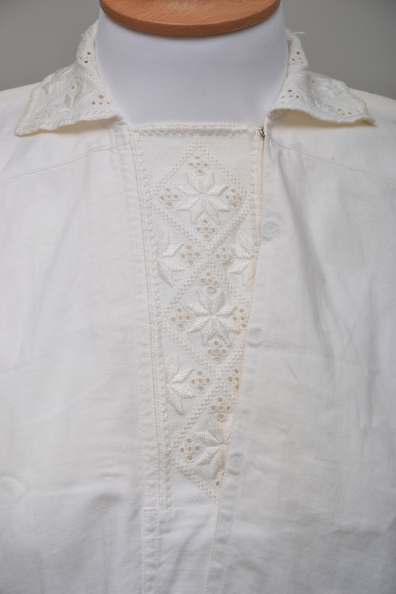 Hvit bomullsskjorte med innfelte stykker med hardangersøm på sultanstoff i stolpe, mansjetter og krage. Lukkes med fire trykknapper i front (to mangler) og en trykkanpp på hver mansjett.