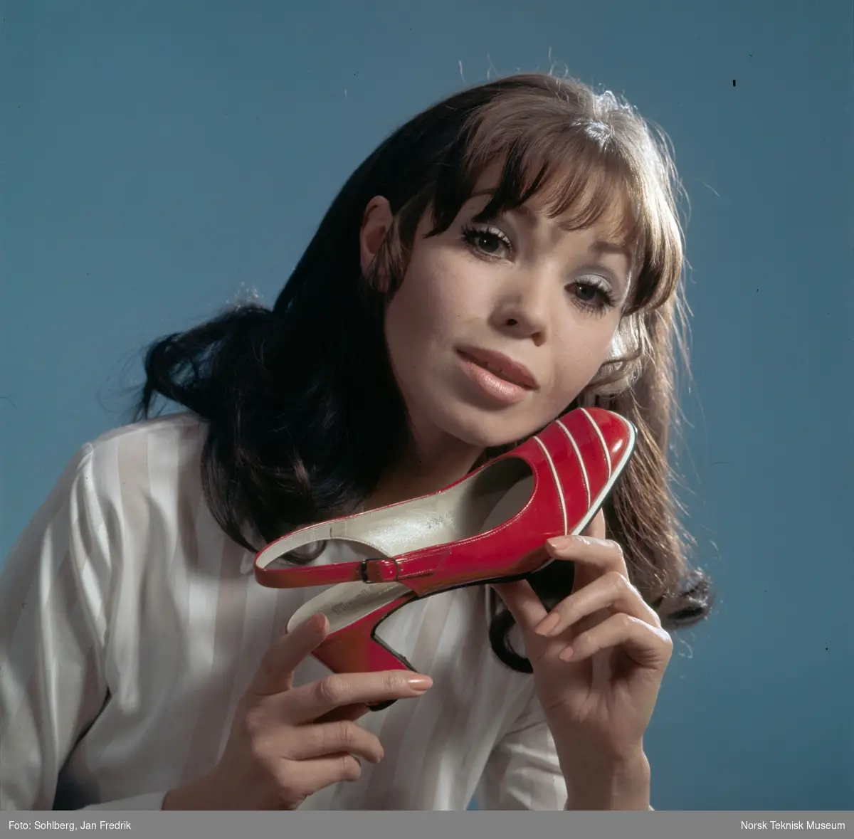 Tidlig norsk reklame- / motefoto. En kvinnelig modell holder en rød sko med hvite striper opp mot kinnet. Hun virker fornøyd med tidens skomote.