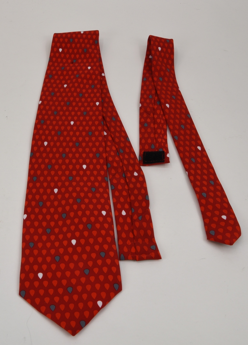 En s. k säkerhetsslips i två delar som sätts samman med hjälp av ett kardborrband i nacken. Slipsen är mörkröd med mönster av ljusare röda droppar samt enstaka vita och grå droppar. Båda delarna är fodrade med en röd textil.