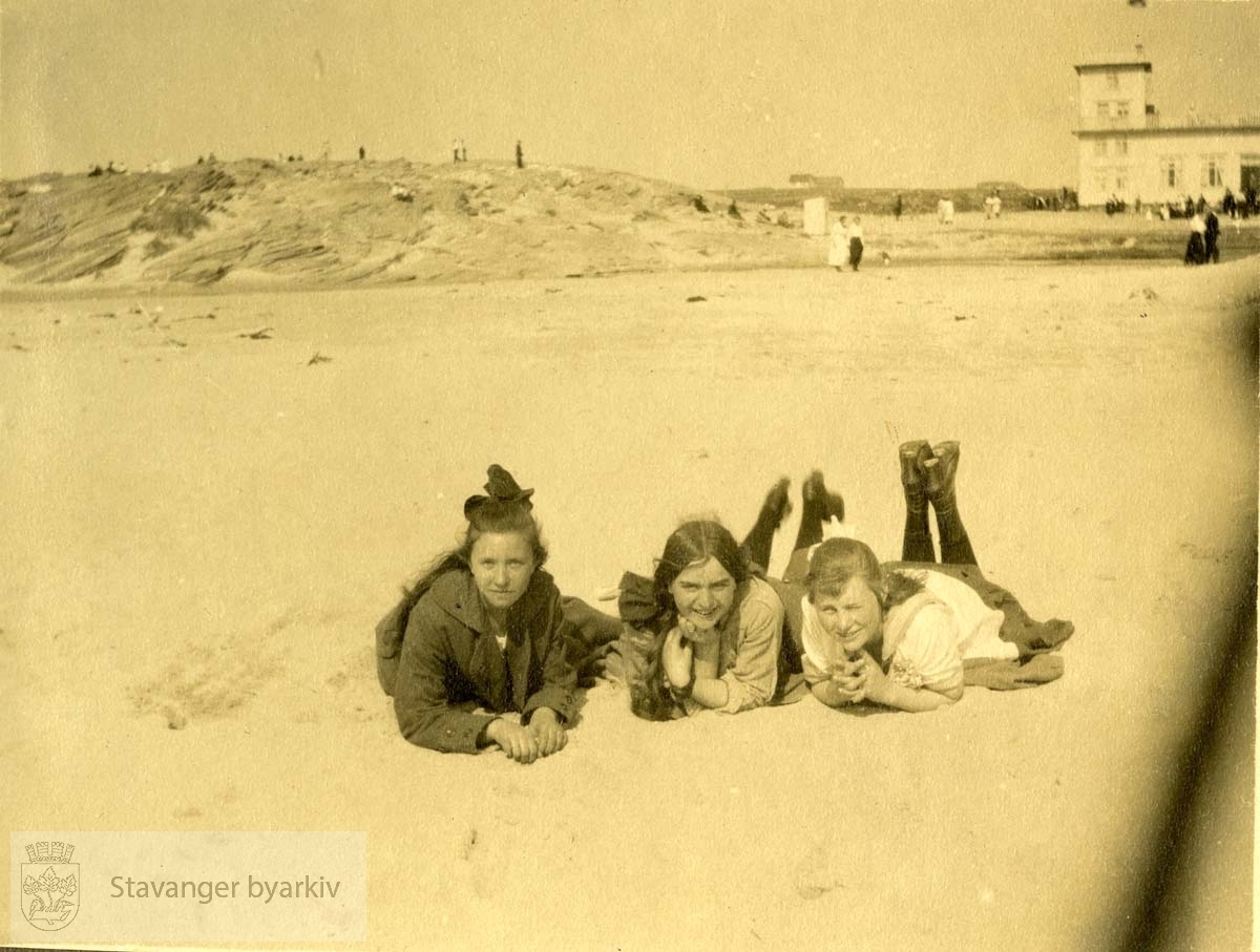 Tre damer ligger på stranden.Sola Strand Hotel i bakgrunnen