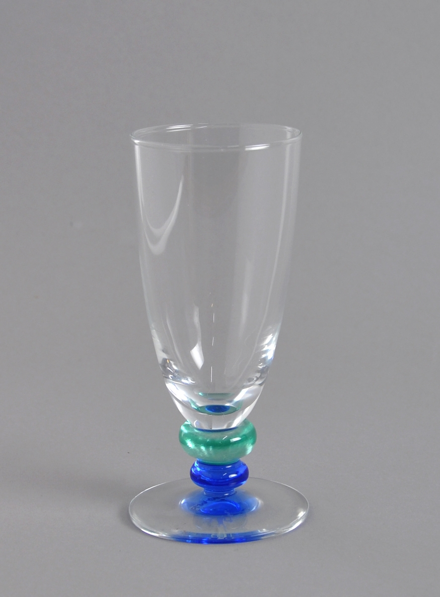Farrisglass med en fakkelmann frostet inn i blått stett. Blå og turkis ring under glasset. Glasset ligger i original emballasje.