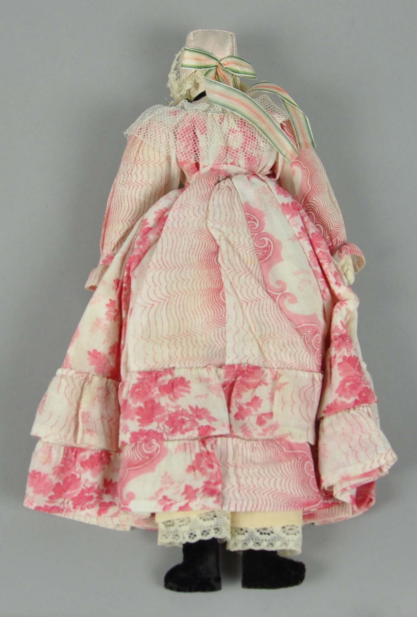 Dokke laget av tekstil. Dokken har lue med blonder på kanten og sløyfe på baksiden og framsiden. Hvit kjole med rosa mønstre og draperinger ved bunnen, og svarte sko.