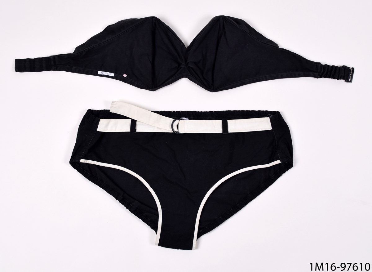 Baddräkt, bikini av svart maskinvävd bomull, med stålfjädrar. Bikini byxorna har ett vitt skärp i midjan samt en vit kant vid grenen framtill.