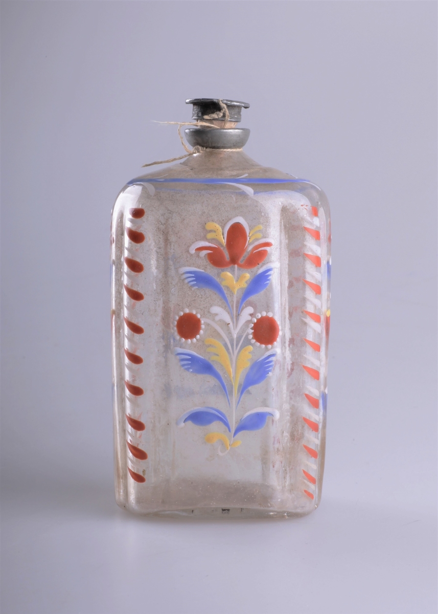 Åttekantet flaske blåst i klart glass. Med tuten innfelt i glassmassen overst, og brutt pontemerke i bunnen. Påmalt med emaljemaling eller blylakk i blått, rødt, hvitt og gult. Dekor i form av stilistiske blomster. Kork og munning i tinn. Kan være av av tysk opprinnelse i tidsepoken 1750-1850.