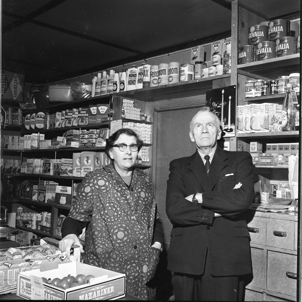 Asta och Olof Landberg bakom disken i sin affär i Ekhagen, Bunnström. I bakgrunden vägghyllor med torrvaror, kaffe, konserver och rengöringsmedel. I förgrunden på disken bland annat en låda märkt Mazariner med tomater