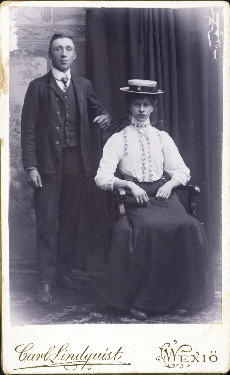 Gruppporträtt av en sittande kvinna i hatt, blus och kjol och en stående man i kostym med väst. 
Helfigur. Ateljéfoto.