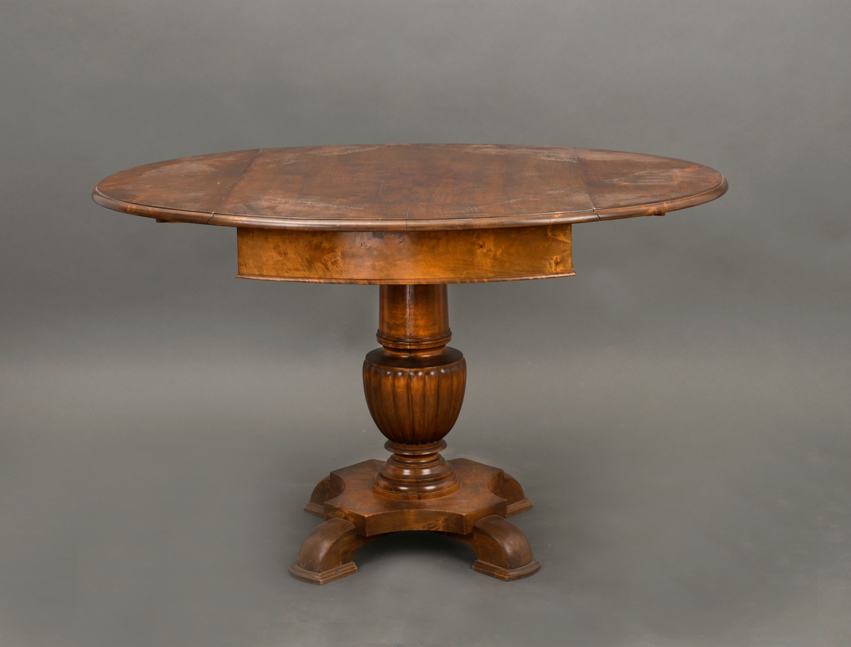 Salongbordet er ovalt og brunt på tykk søyle som har fire føtter. Bordet har to klaffer.