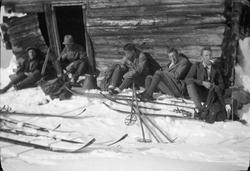 Fem menn sitter langs husvegg og tar en pause fra skitur