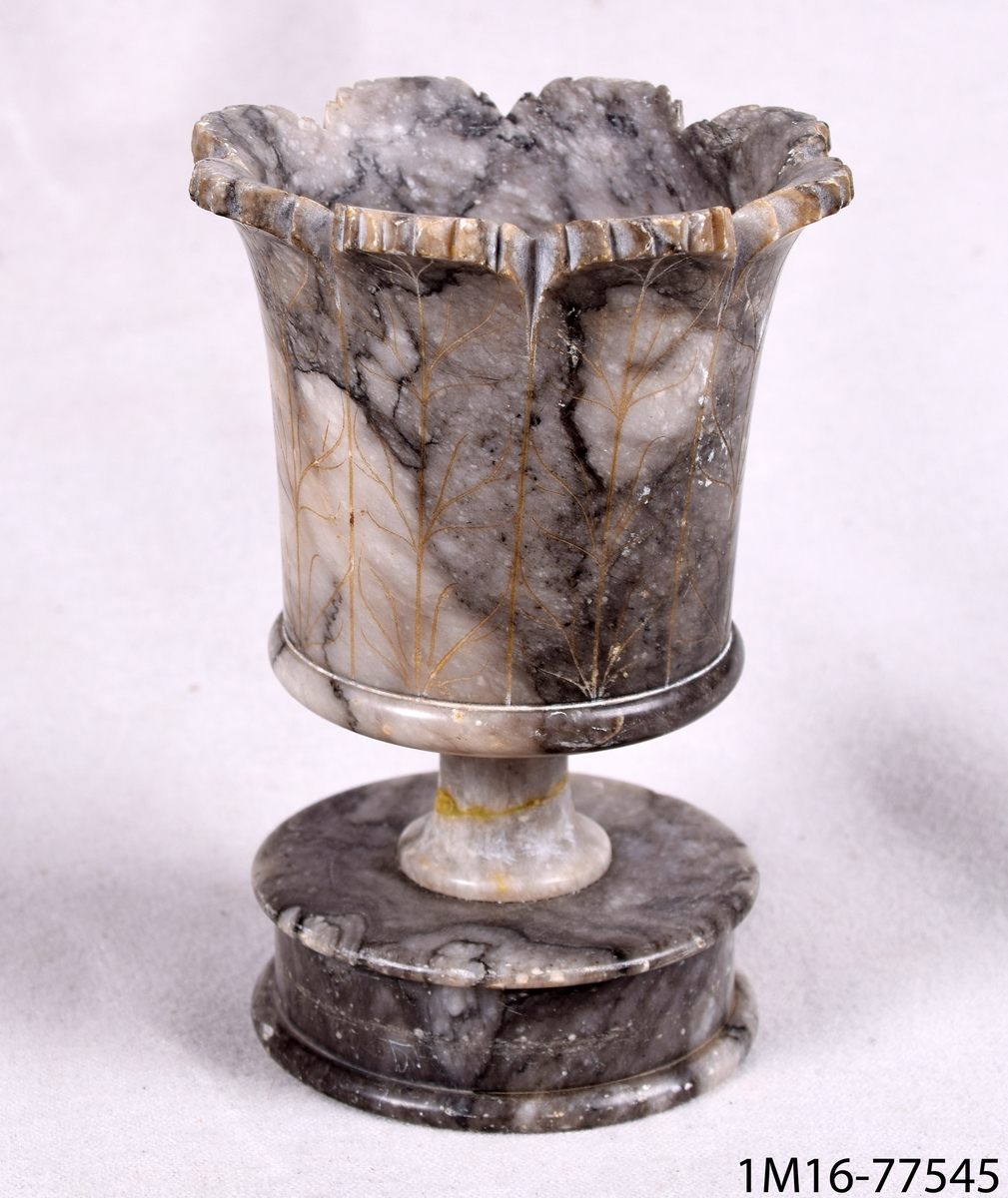 Cigarrställ av gråflammig marmor. Stället har en rund fot och i övre kant, vid mynningen, flikig blombladsliknande dekor.