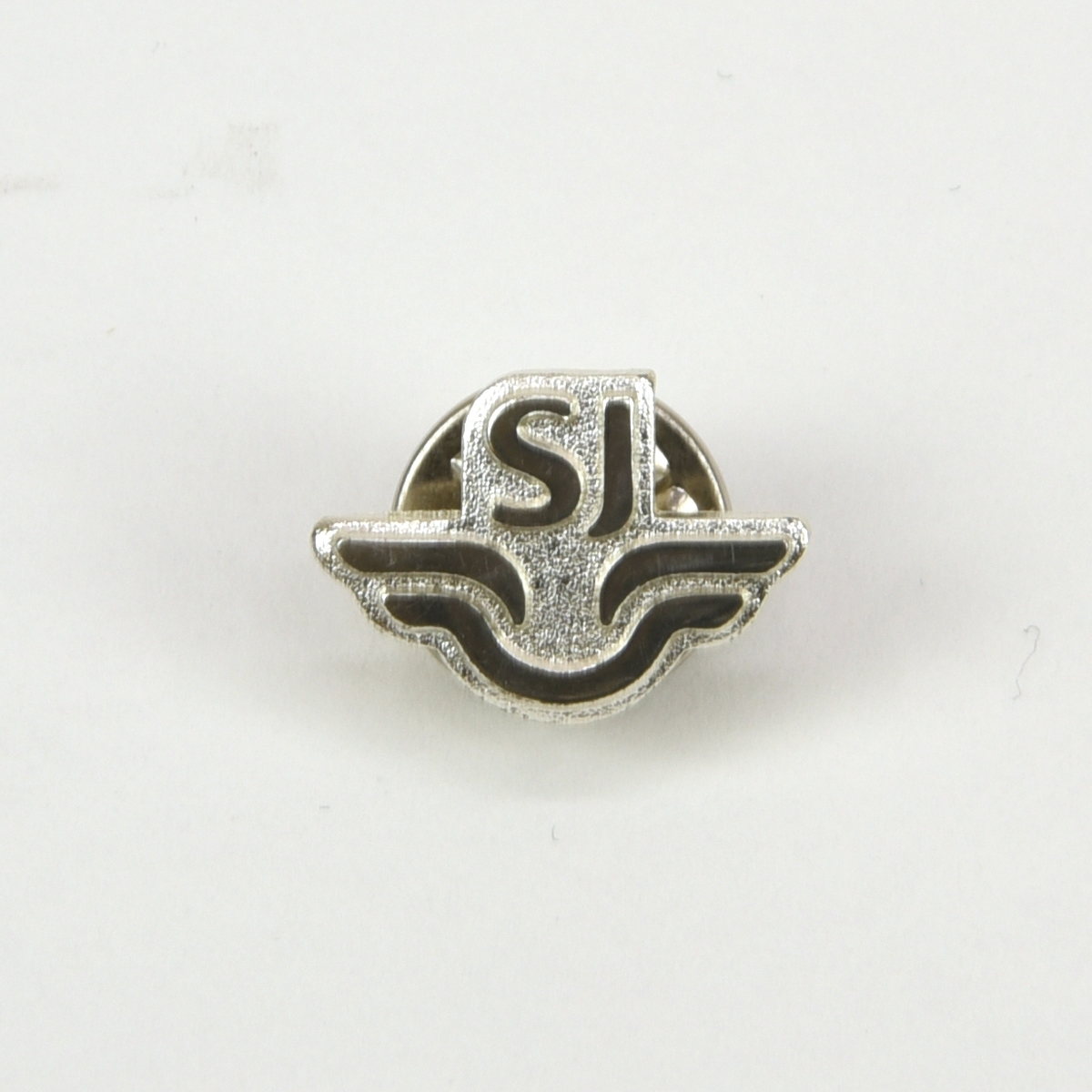 Liten metallnål med SJ:s logotyp i form av ett stiliserat vingpar med "SJ" ovanför. På baksidan finns en nål med ett runt lås.