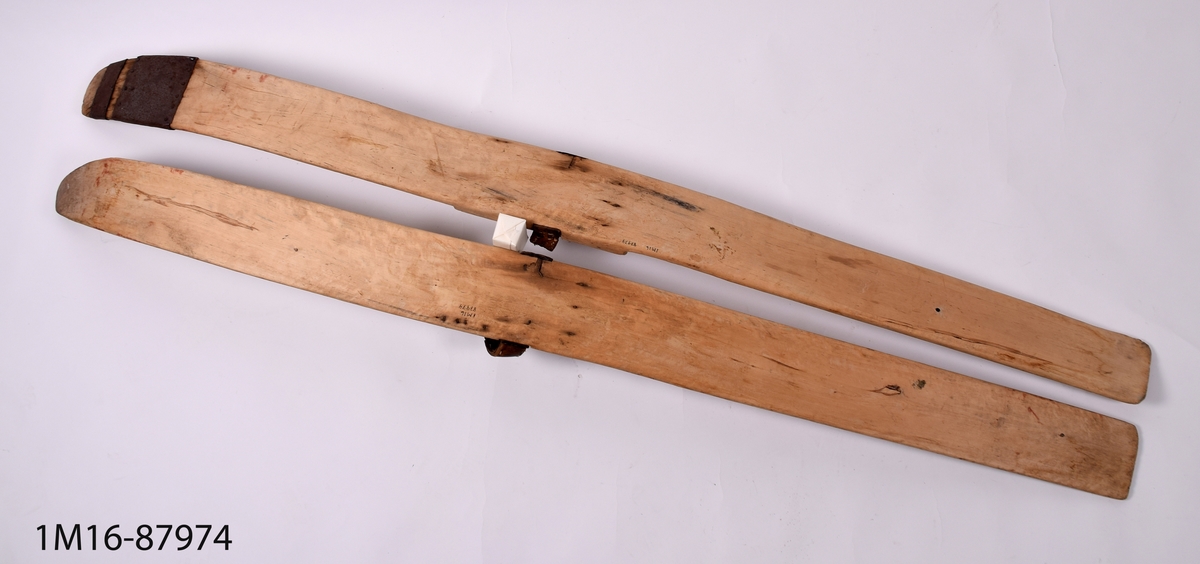 Skidor, ett par, av lövträ med läderbindsrem. Den ena skidan lagad med järnplåt.
