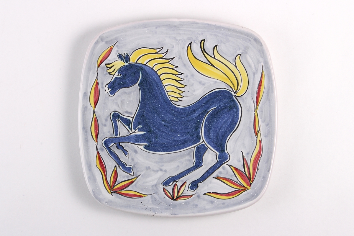 Firkantet tallerken dekorert med en blå hest som har gul man. Med hull til oppheng på baksiden.