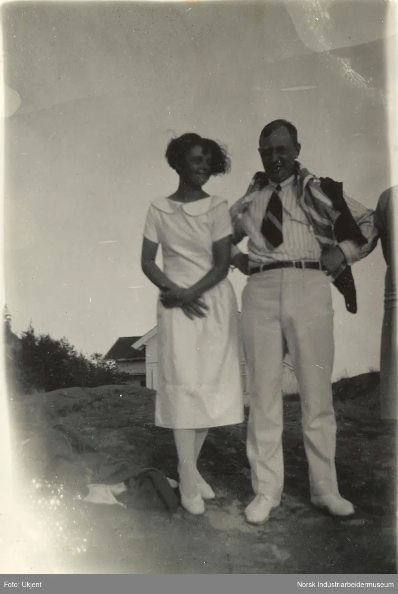 Mann og jente i lyse sommerklær står på svaberg foran sommerhytte