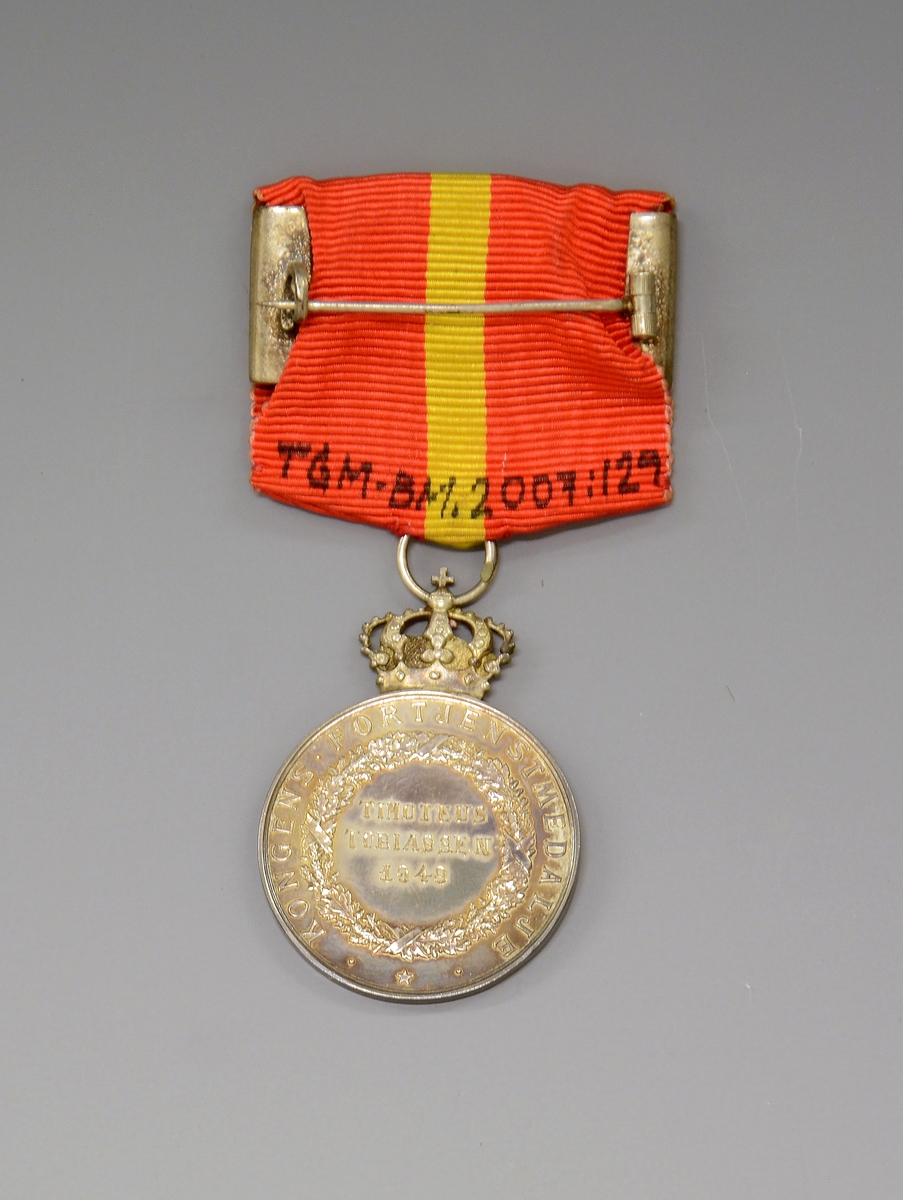 Medaljen ligger i en liten, flat, burgunderrød eske (original-etui) - preget med en forgylt krone på lokket. Silke og fløyel innvendig.