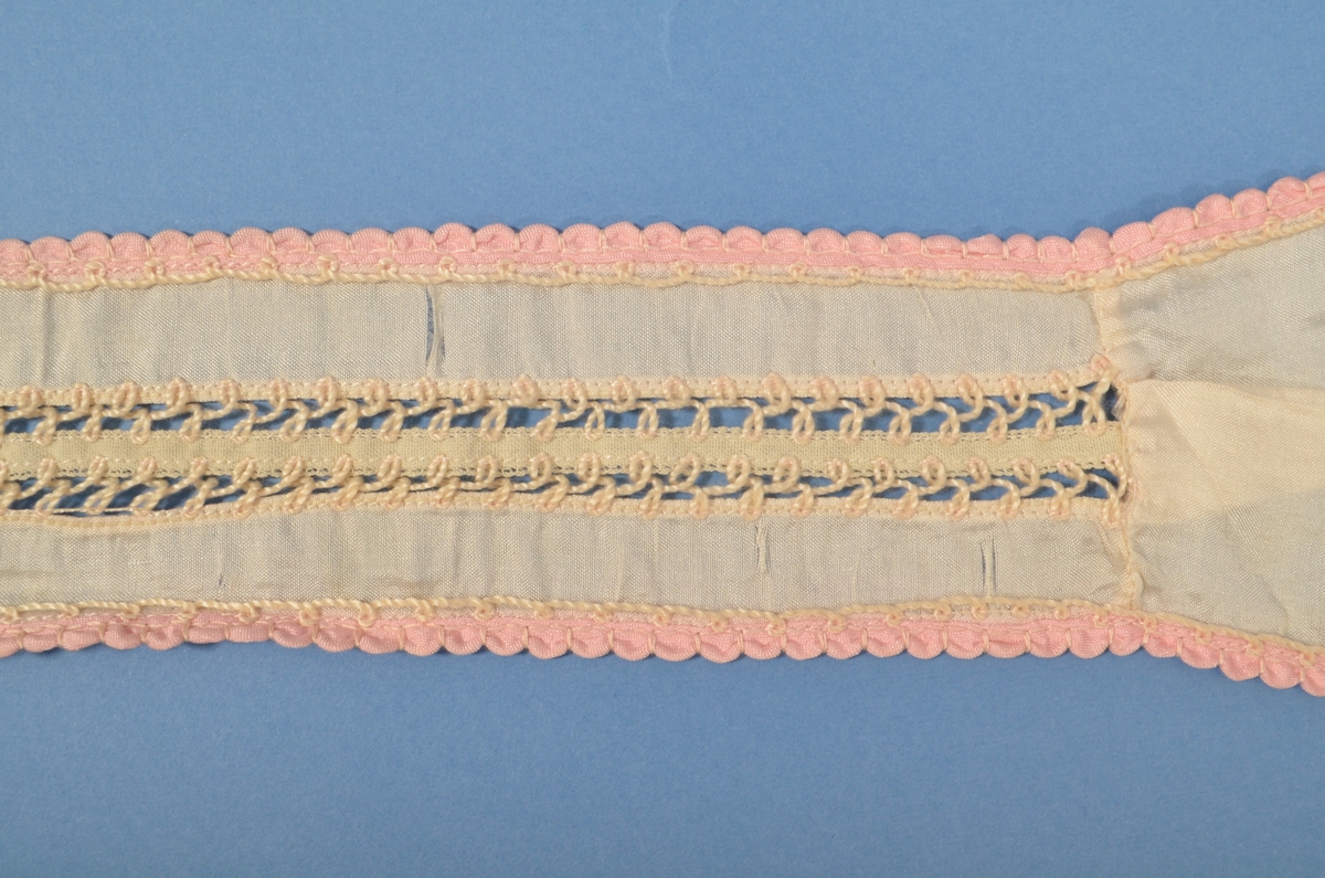 Halssløyfe i tynn kvit silke, kanta med rosa band. Midt på bandet pryda med band og kryssesting.