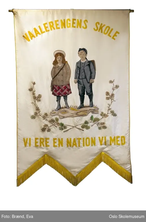 To barn med ransel på lys bakgrunn. Øverst på fanen står skolens navn. Nederst på fanen står teksten "Vi ere en nation vi med!" som er første linje fra en barnesang, egentlig "Smaaguttenes Nationalsang" av Henrik Wergeland.