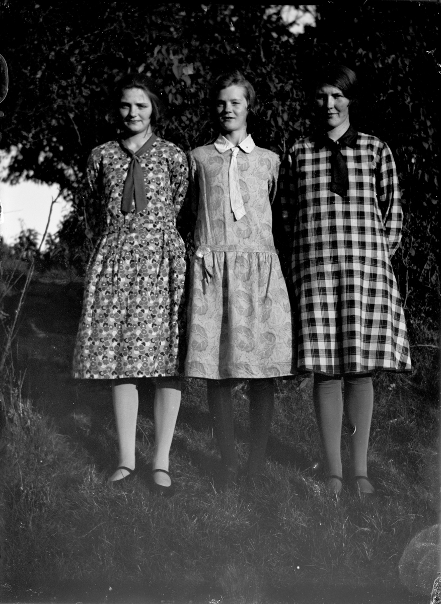 Tre äldre flickor poserar i gröngräset i finklänningar av 1930-talssnitt med slipsar.