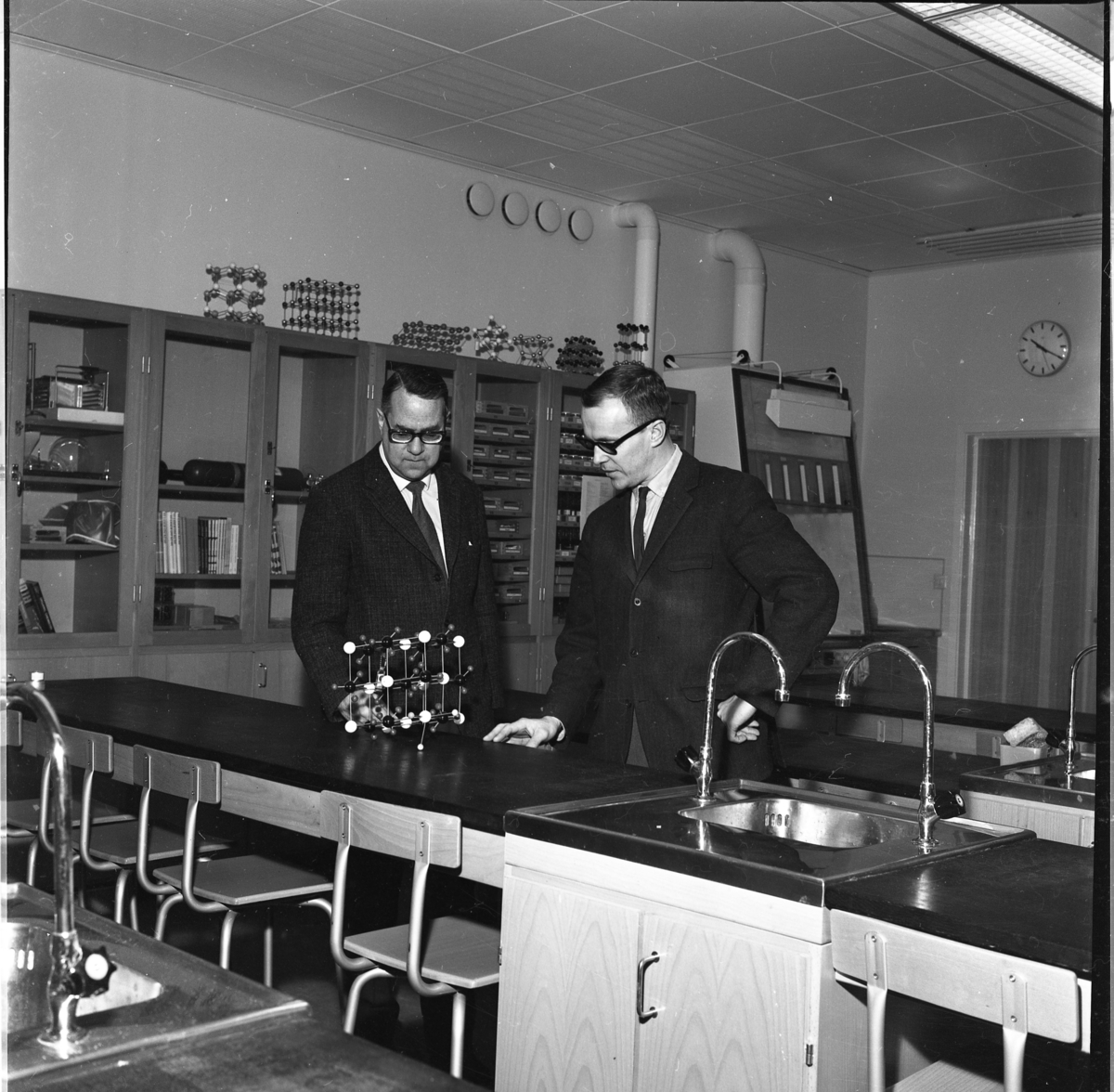 Två män står vid en lång skolbänk som separeras av ett tvättställ. De står framför en modell av en molekyl. Utmed väggen i bakgrunden står höga skåp med glasdörrar. Det är rektor Göte Östlund och ämneslärare Hans Fridell till höger som befinner sig i en lärosal på Ribbaskolan.