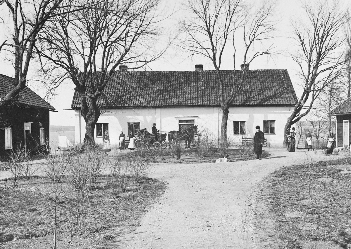 Någon gång under 1870-talets första hälft förevigade den danskättade fotografen Peder Anton Eriksén denna vy mot Ekebyborna kyrkoherdeboställe. Vackert belägen på en udde vid sjön Boren. Ursprungligen uppförd 1735 och länge ansedd som en av de "vackraste och bästa" i Aska kontrakt. Långt senare var bygganden dock i dåligt skick och 1928 byggdes ny prästgård närmare kyrkan. Året därpå skänktes den gamla prästgården till Nordiska museet för att flyttas till Skansen som prästgård till Seglora kyrka. Planen blev emellertid inte av utan huset blev kvar på ursprunglig plats och hyrdes ut. År 1989 skänkte Nordiska museet prästgården till Ask-Ekebyborna hembygdsförening. Samtidigt förklarades den som statligt byggnadsminne. Vid tiden för bilden disponerades bostället av kyrkoherde Per Dahlgren med familj. Det är honom vi ser intill rundeln till höger, rimligtvis vid sidan av maka, barn och tjänstefolk.