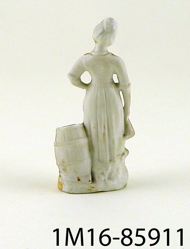 Figurin föreställande kvinna i vitt med hatt och rött livstycke, vit blus och vit uppskörtad kjol med grön underkjol, hållande ett krus i handen stående vid en ihålig tunna. Tunnan kan användas som vas.