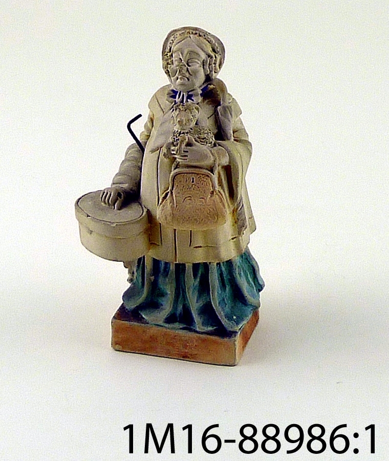 Figurin föreställande kvinna som bär på korg, väska, paraply och liten hund. Figurinens utförande har drag av karikatyr.