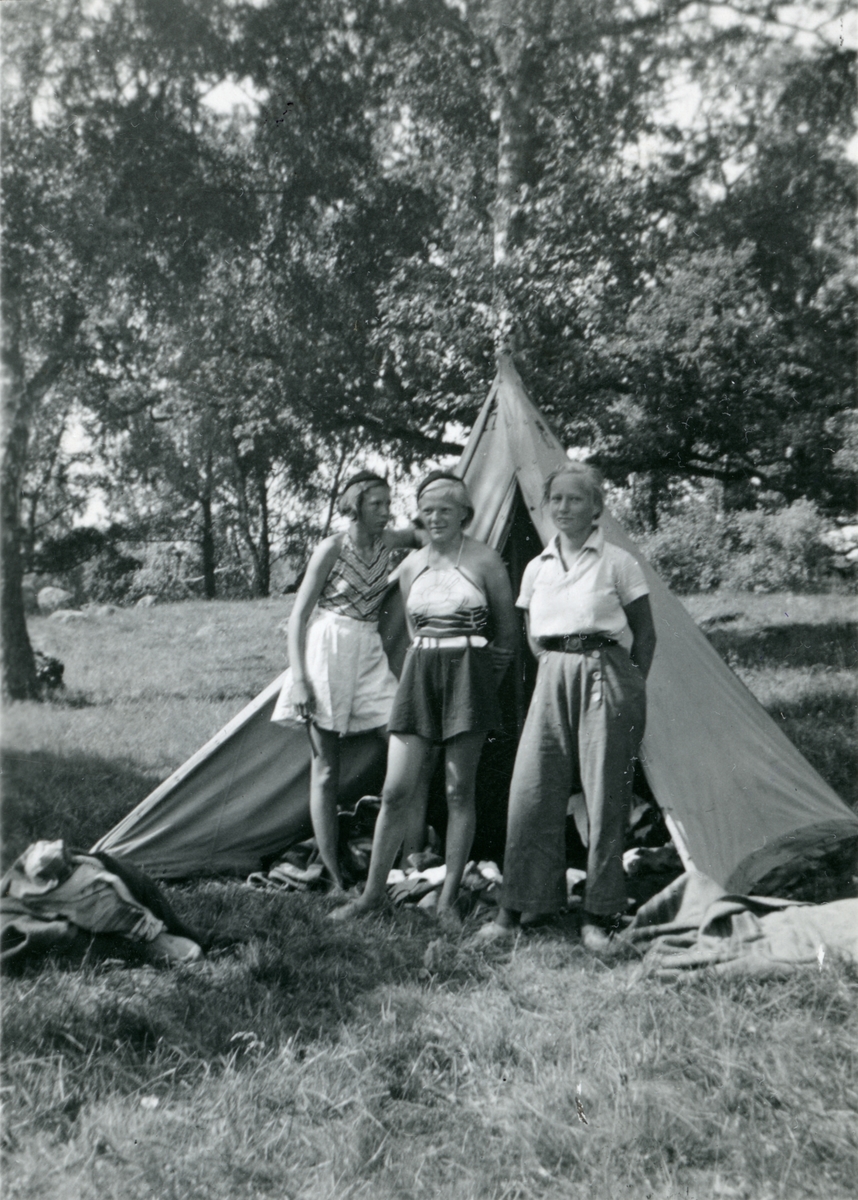 Tre unga kvinnor framför ett tält i naturen.