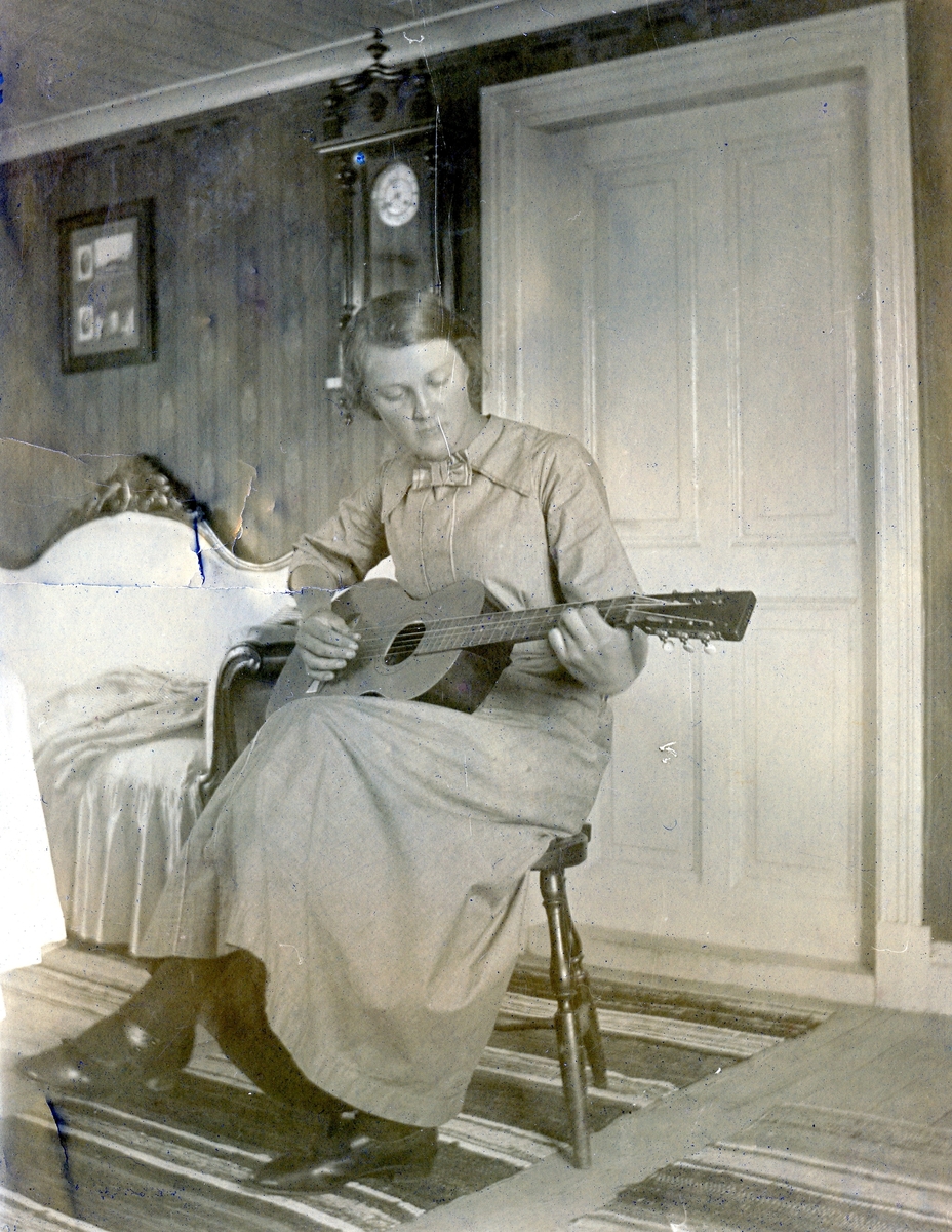Ester Jakobsson (1896-1926) sitter på en stol och spelar gitarr inne i boningshuset Lilla Vommedal (idag: Streteredsvägen 25) 1913-08-10. Ester var fosterdotter till Anton och Maria Gustafsson, ägare av Lilla Vommedal. Hon var också syster till Oscar Jakobsson (1889-1945), skaparen av den folkkäre seriefiguren Adamson*. 
Esther avled i TBC 1926.