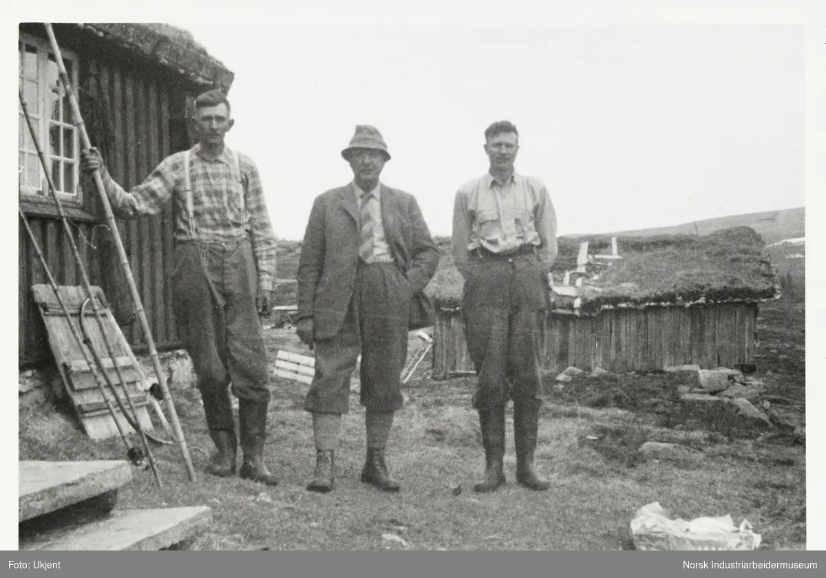 Tre menn står på gårdstun ved hytte. Fiskestenger plassert langs vegg