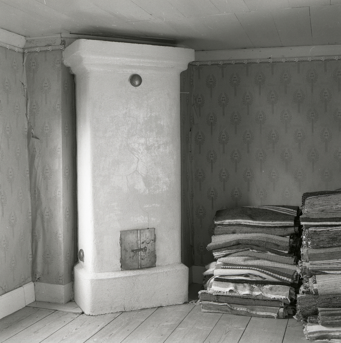 I ett rum står en vit kakelugn och bredvid ligger det trasmattor i travar. På väggarna är det gamla tapeter och det är trägolv och trätak. Utegårn 19 augusti 1990.