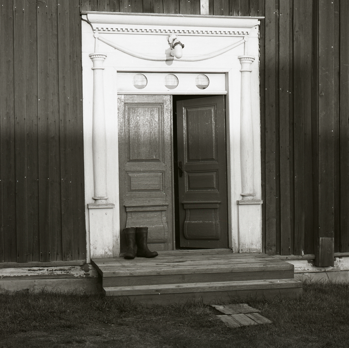 Ett par stövlar står på en trappa utanför en dörr. Dörren smyckad med snidad dekoration föreställande kolonner och girland, Boteå 1974.