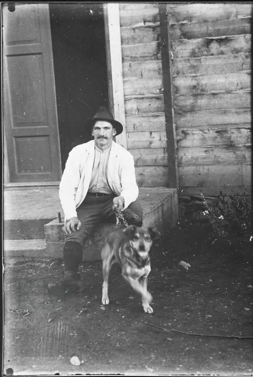 En mann sitter på ei trapp utenfor inngangen til et hus. Han holder en hund i bånd.