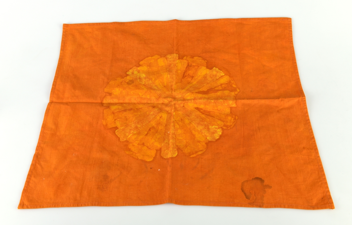 En bordduk laget av bomullsstoff. Den er firkantet og falden er maskinsydd. Duken er oransje med mønster i oransjegult og gult. Mønsteret er minner om en sol eller en stor blomst. Mønsteret er på midten av duken og er trolig laget ved batikk.