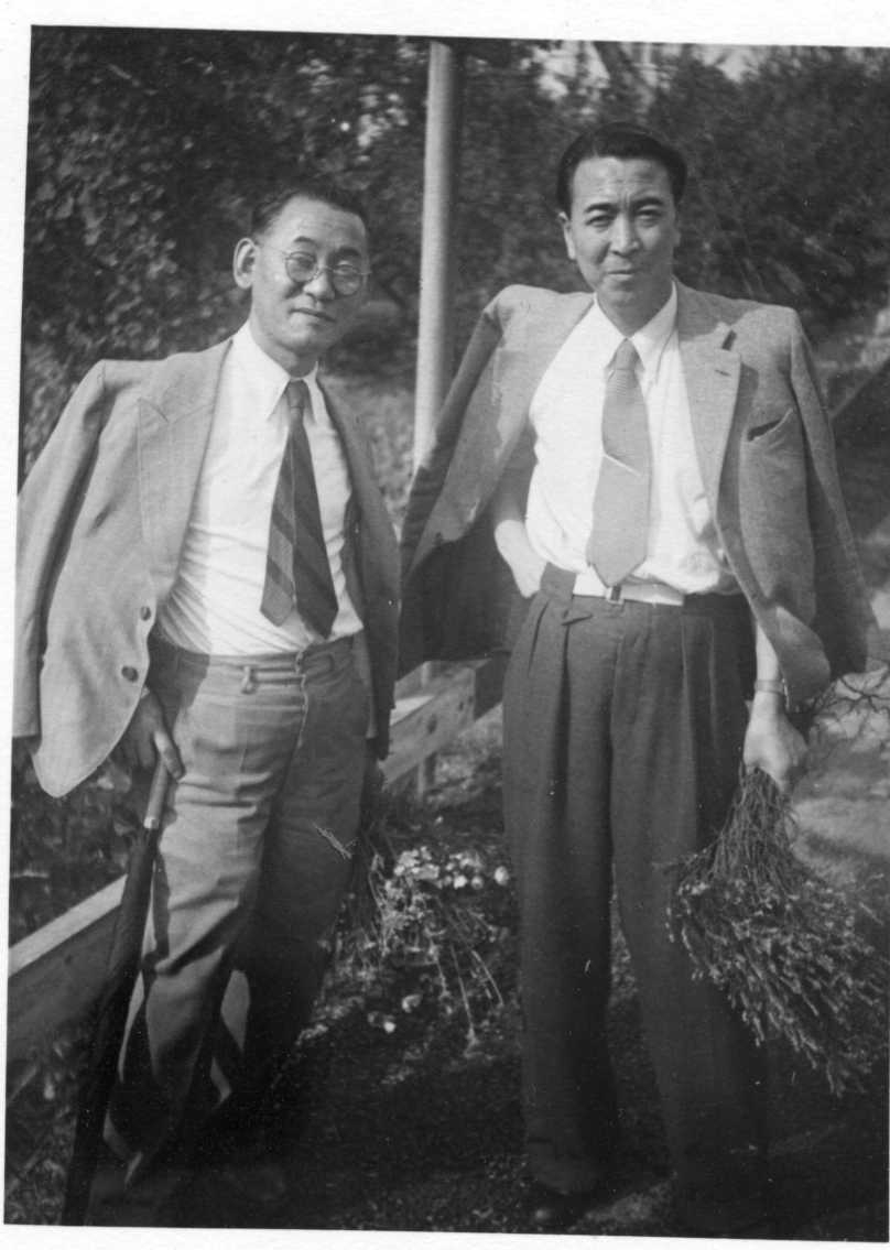 Två välklädda japaner i slips med kavajen hängande över axlarna. En har ett ihopfällt paraply i handen. Bägge har en bukett med ängsblommor i handen.