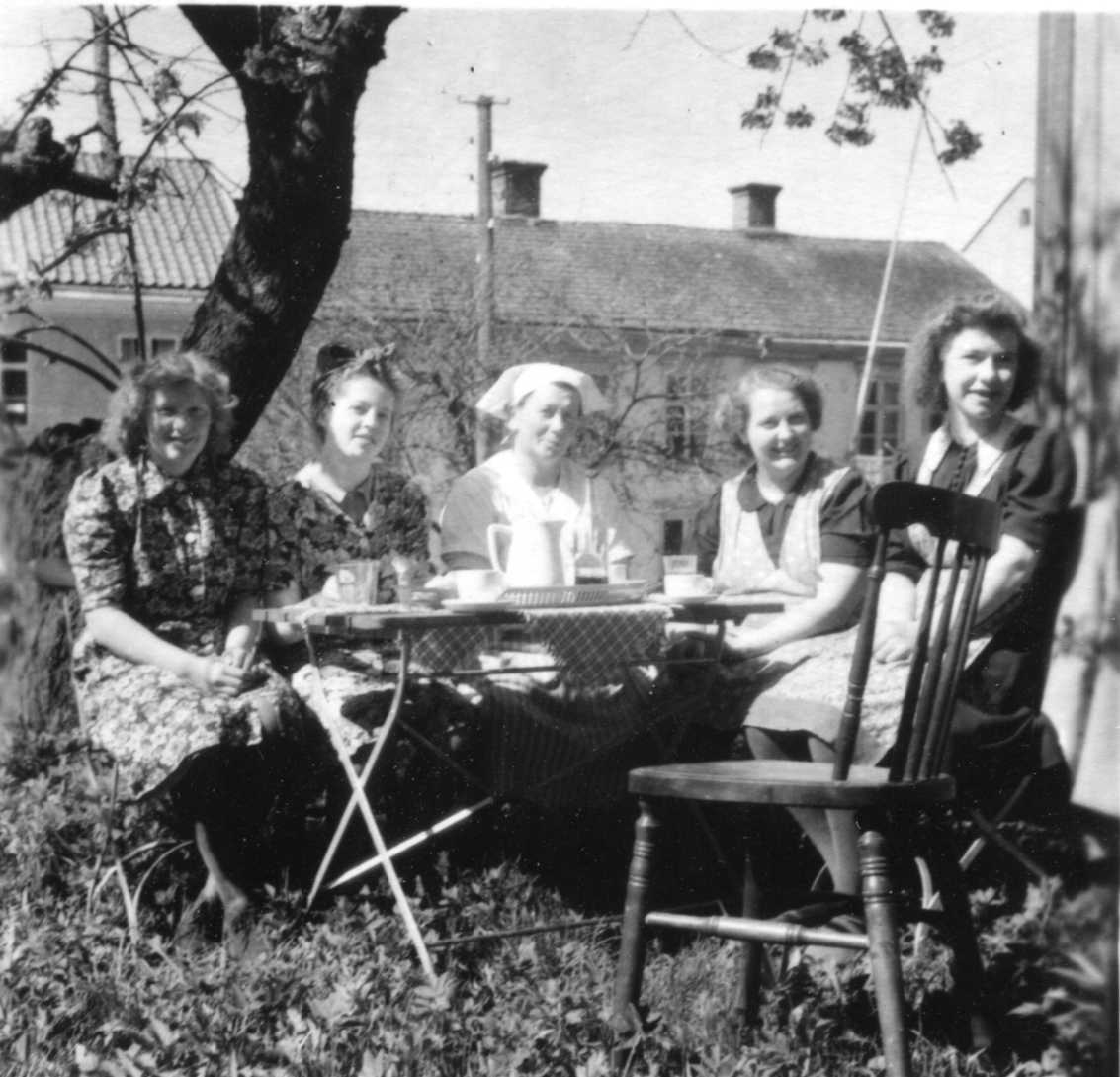 Fem kvinnor sitter i en trädgård vid Brahegatan runt ett bord med kaffekoppar. Två kvinnor till vänster bär blommig klänning. Kvinnan i mitten är klädd som kokerska och de två till höger likt servitriser. Kokerskan heter Anna Andersson.