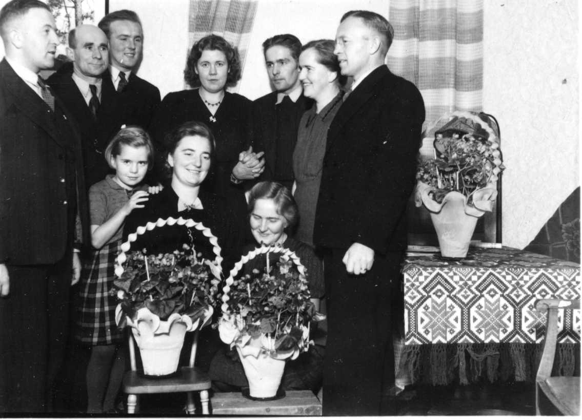 Gruppbild med släkterna Simonsson och Granstrand. På en stol respektive pall framför dem står två blomsterkorgar. En liten flicka håller händerna på en kvinnans axel.