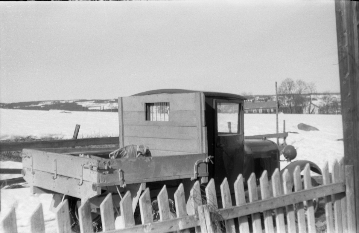 Snødekt landskap med en liten lastebil i forgrunnen. Midt i bildet bygninger på gården Skjefstad i Lensbygda. Fotografens standplass har trolig vært ved sveiserboligen på gården Nordre Rise. Bilen skal iflg. informant være Ford pickup av årsmodell 1928-29.
