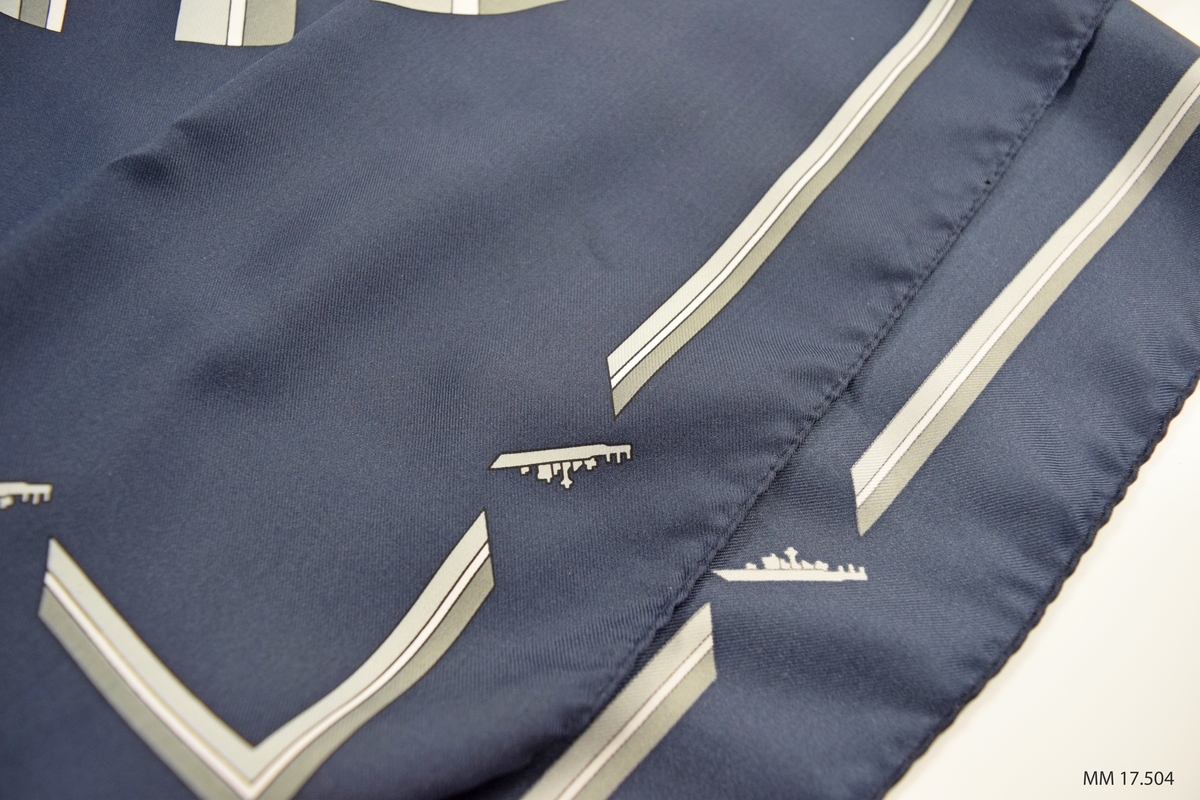 Marinblå scarf med tryckt mönster, grå och vita diagonalt ställda breda ränder lagda i cirkelformat mönster i mitten. Längs kanten rand avbruten på båda sidor om varje hörn av små fartyg, kustkorvett typ Stockholm. I ena hörnan vit etikett, angivande kvalitet och tvättråd.