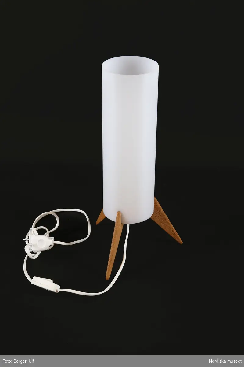 a-d) Bordslampa, elektrisk. b) Skärm av vertikalt rör av vit akrylplast på a) tre spetsiga fötter av ljus ek. c) Matt glödlampa och d) vit plastsladd med stickpropp.

Den vita sladden med jordad kontakt monterades i lampan i samband med utställningen Nordiskt ljus 2016. Den vita originalsladden, förlängd och skarvad med en så kallad sockerbit, och med ojordad stickpropp, är bevarad.
/Maria Maxén 2017-06-26
