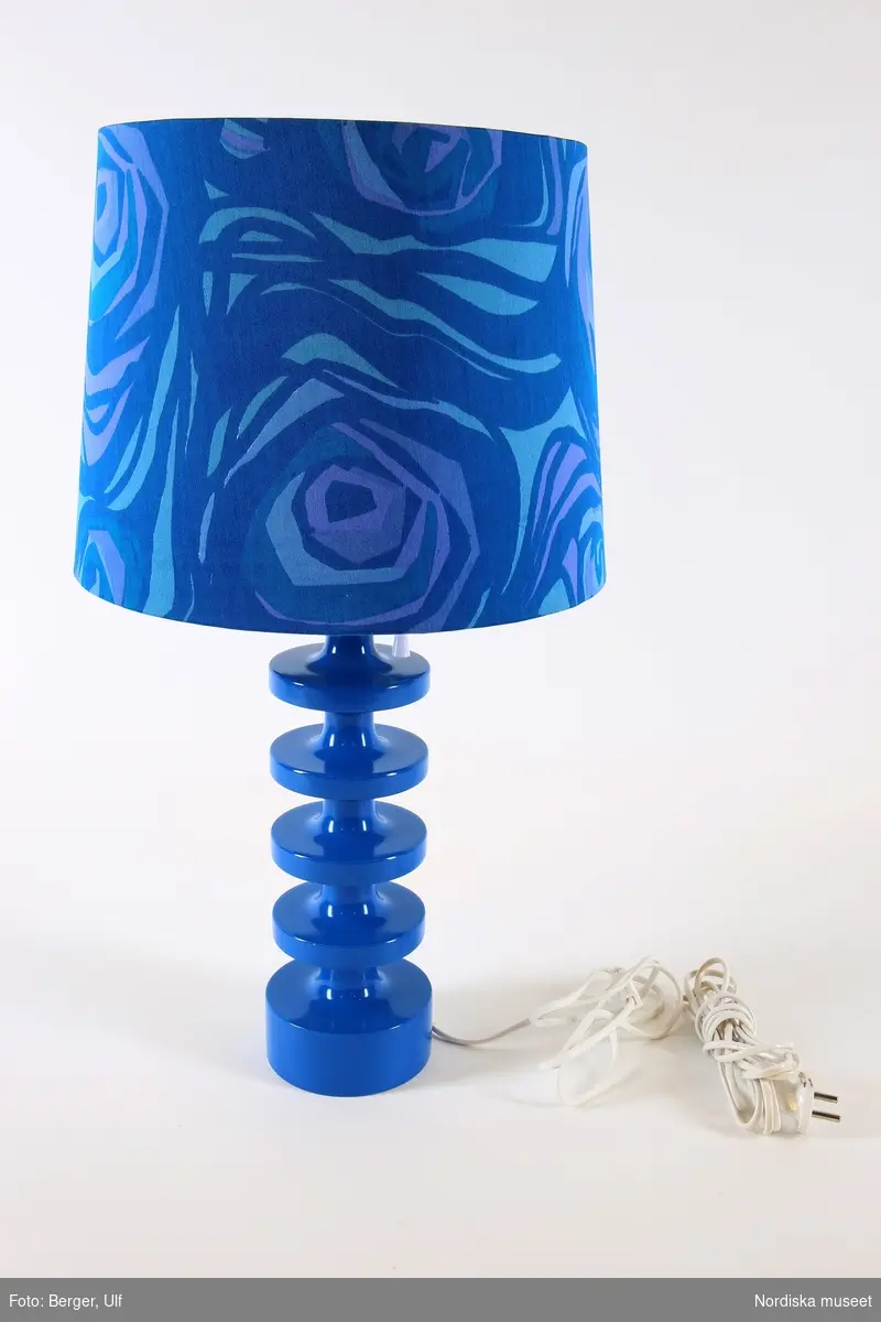 Bordslampa, a) cylindrisk fot med fyra noder, målad i högblank blå färg. Högst upp på foten strömbrytare i form av vitt dragsnöre. b) Närmast cylindrisk lampskärm av plast klädd med mönstrat tyg i blått. c) Matt glödlampa, d) vit plastsladd med ojordad kontakt. Den isittande sladden monterades i lampan i samband med utställningen Nordiskt ljus 2016. Originalsladden, vit med ojordad stickpropp, bevarad.

Texolux-skärm i plast där mitten av skärmens opalvita "tak" bildar en nedåtgånede strut som omsluter glödlampan, för ett bländfritt ljus.
/Maria Maxén 2017-06-26