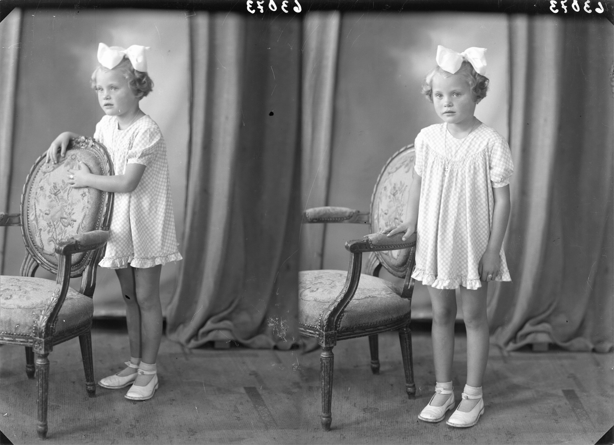 Portrett. Ung lyshåret pike i kort lys smårutet kjole og lys sløyfe i håret poserer sammen med en brodert stol. Bestillt av Liken Hansen. Salhusveien.